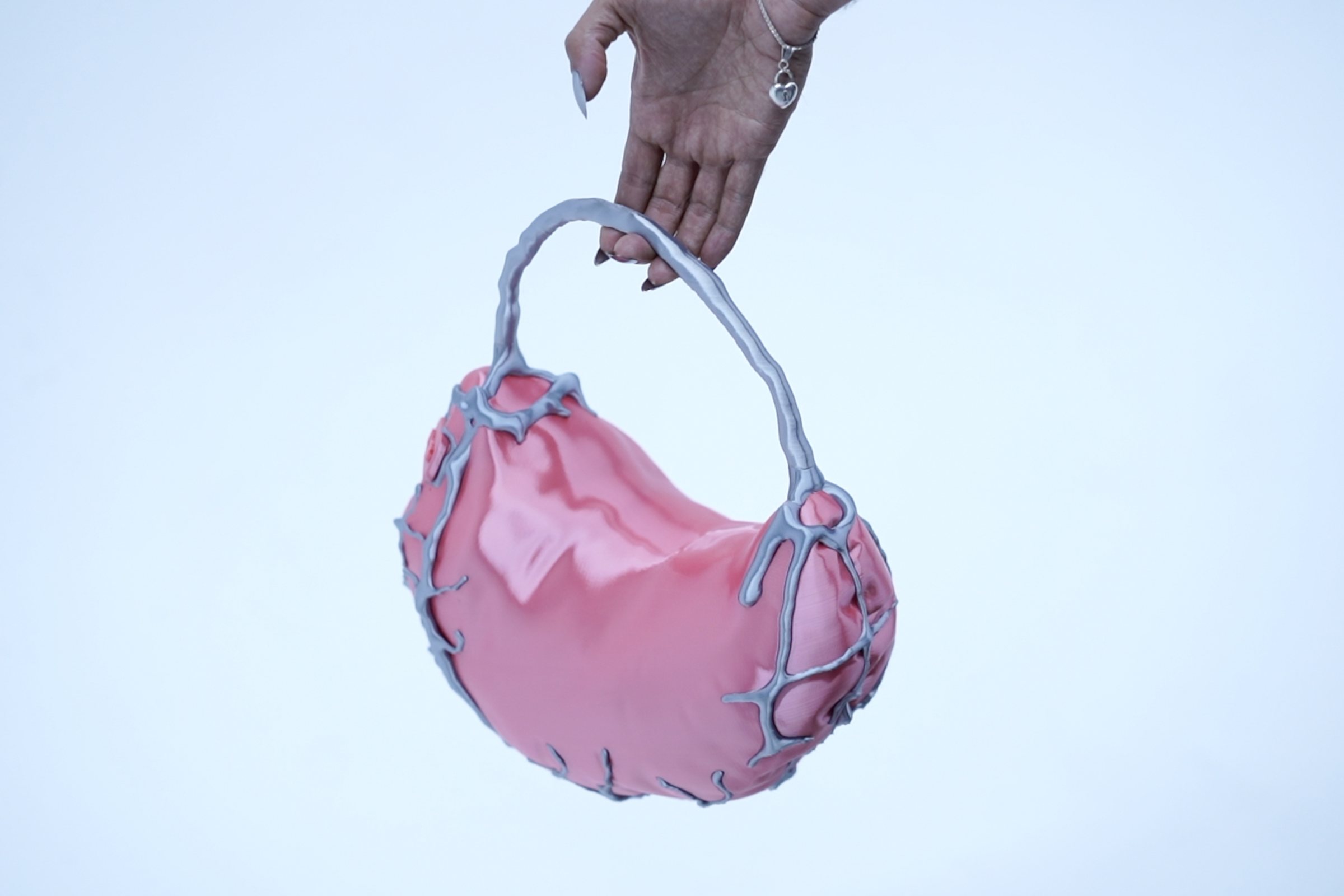 A hand with long nails holds up Ecto Bag, a glossy pink womb-shaped handbag veined with silver, against a pale blue sky