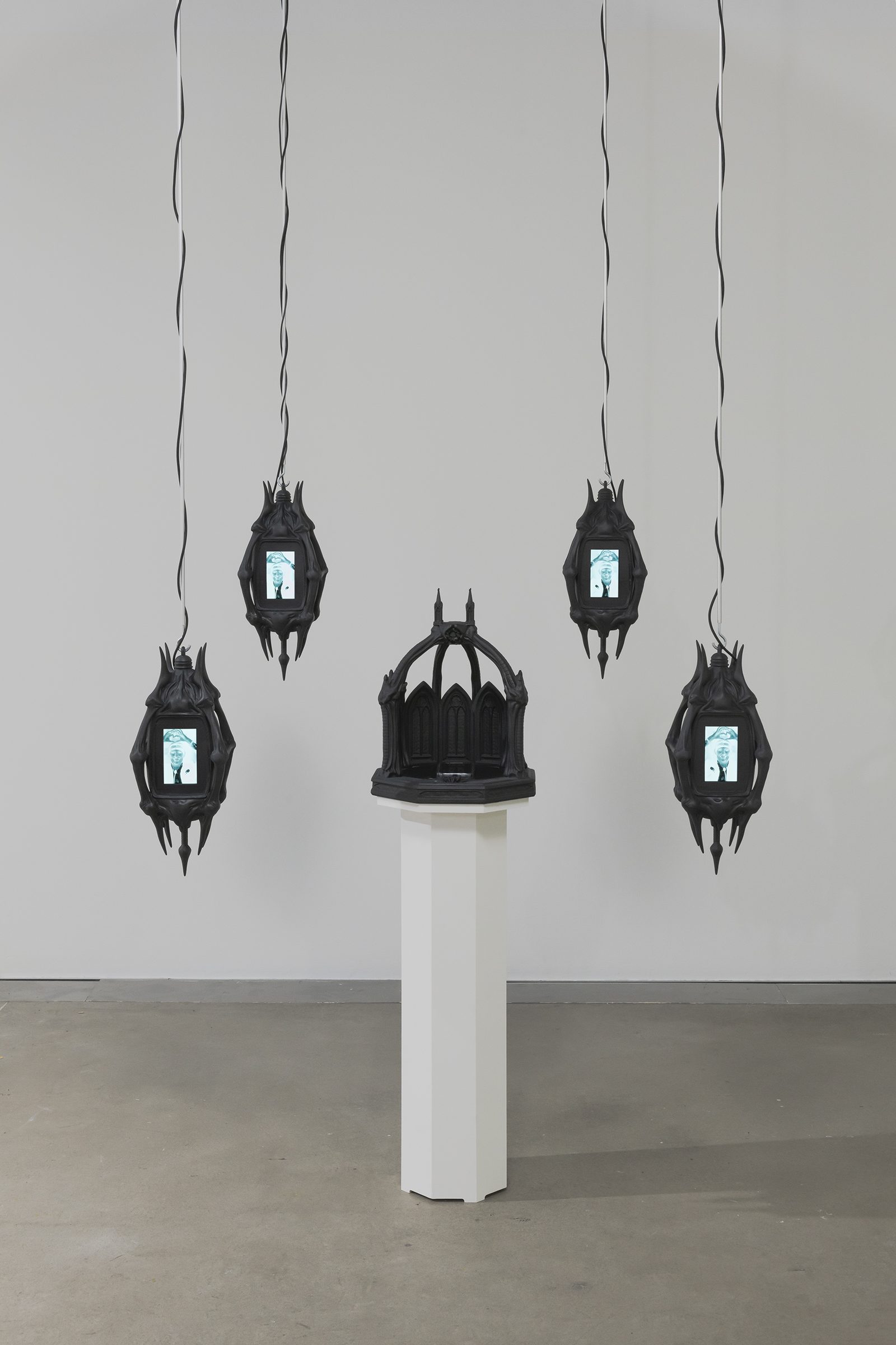 Four black gothic reliquaries with small screens hang from cables around a black chapel-like shrine on a white plinth