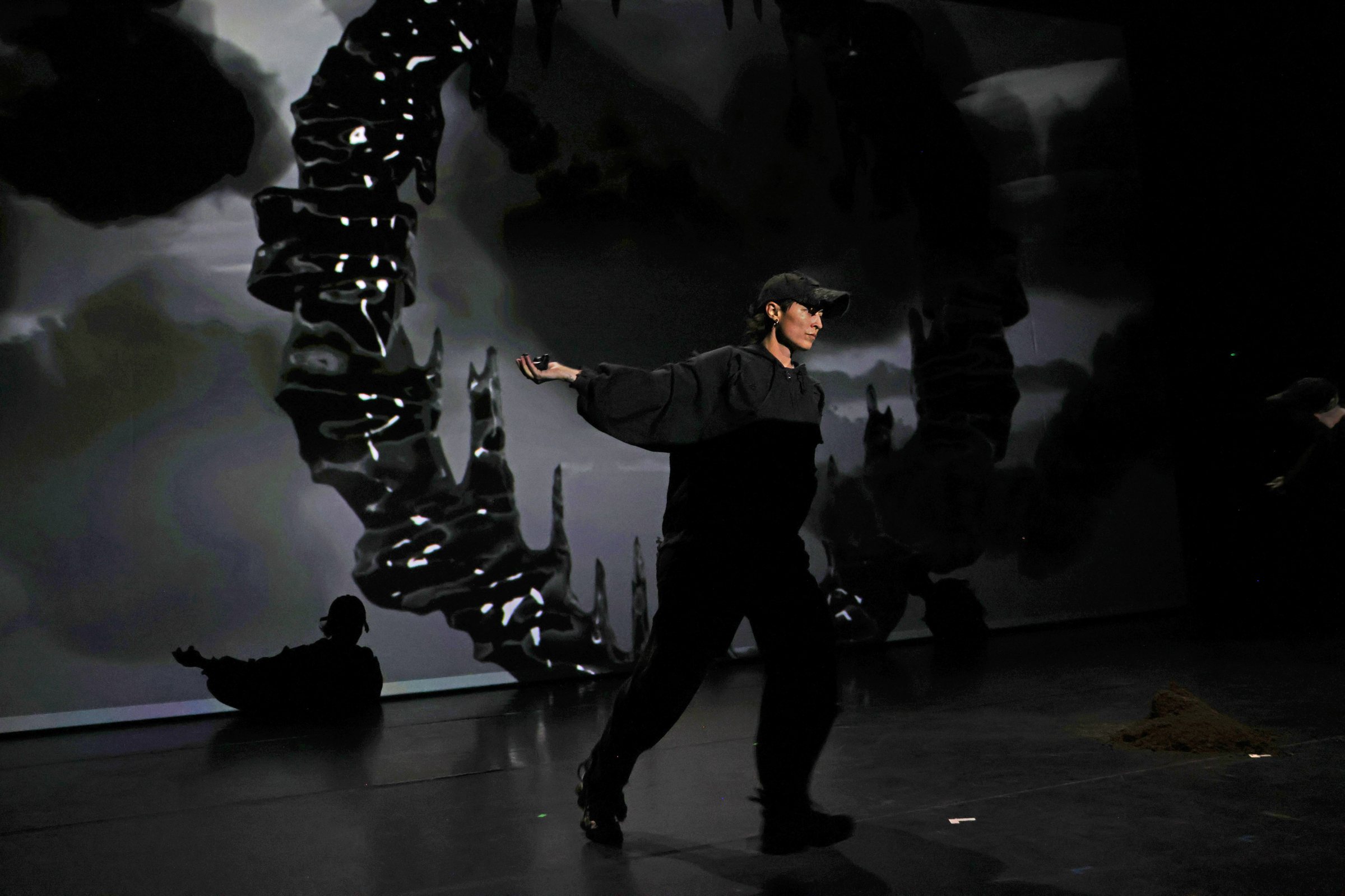 A performer in black with a cap stretches one arm out in front of a projected dark, glistening arch, a second performer seated in the background