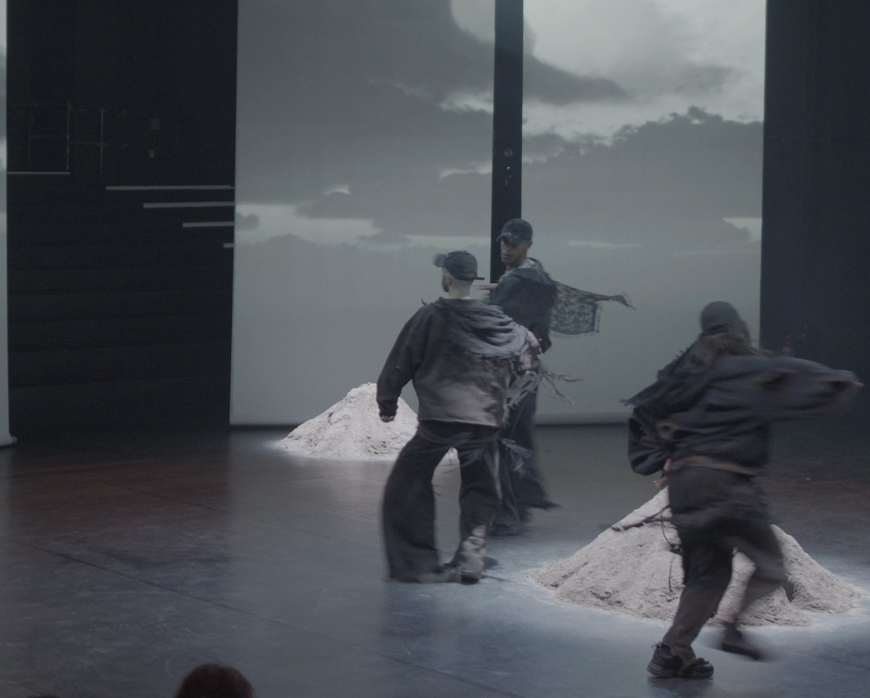 Three performers in layered grey rags move between mounds of pale earth in front of projected cloudy skies, audience in the foreground