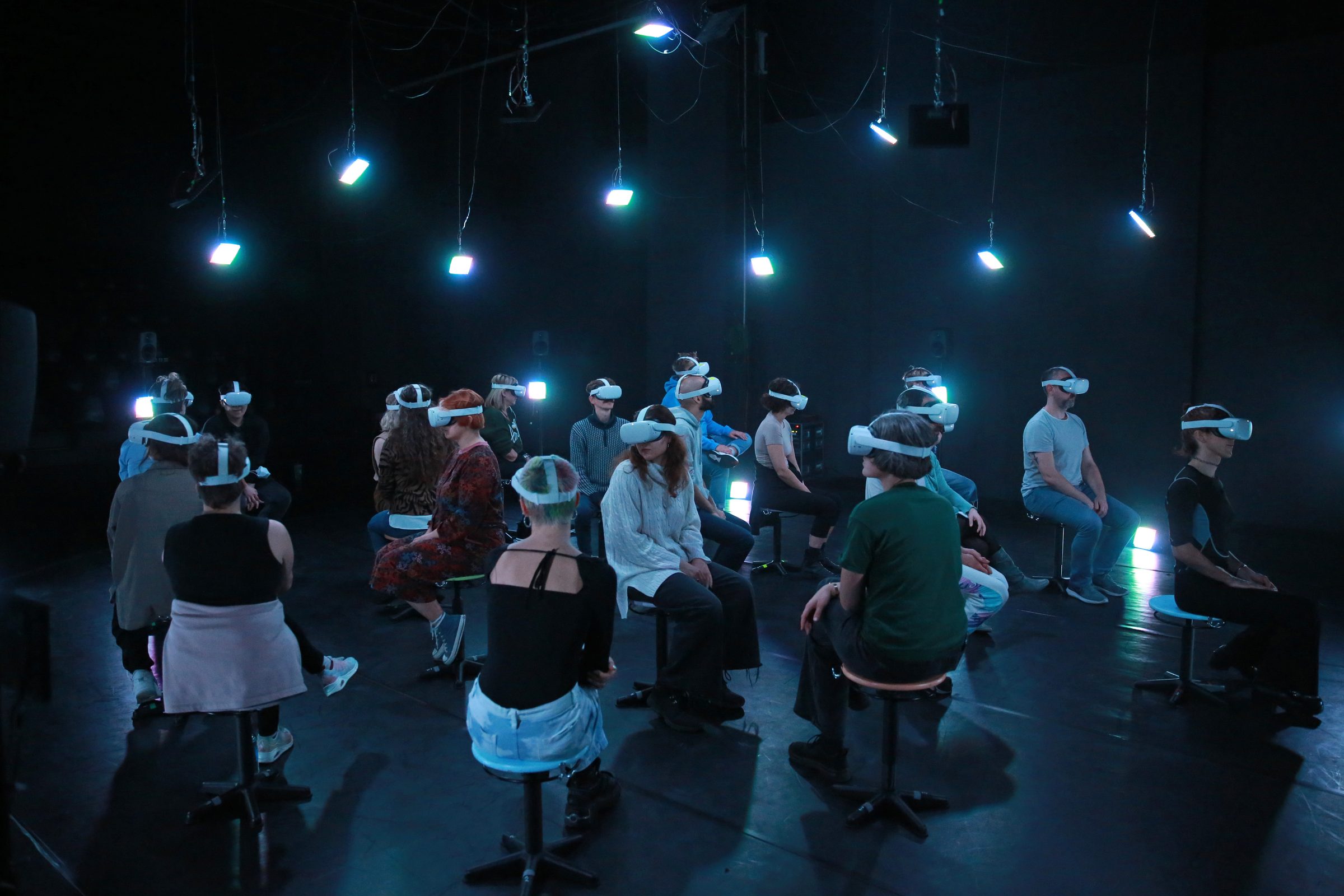 An audience wearing white VR headsets sits on stools in a dark room under hanging blue-green light panels